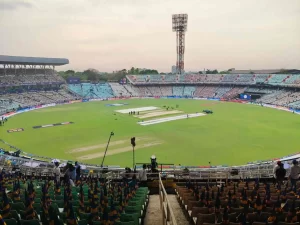 Eden Gardens cricket stadium in Kolkata showing the iconic ground with prepared pitch, floodlight tower, stadium seating sections with green and brown chairs, ground staff preparing covers, and spectators gathering before match - illustrating the historic venue's capacity of 63,000 and balanced black-soil pitch conditions for IPL, T20 World Cup, and international cricket matches