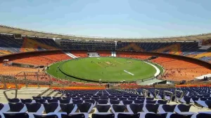 Aerial view of Narendra Modi Stadium in Ahmedabad, India, showing the vast green cricket pitch surrounded by iconic orange and blue seating stands under a clear blue sky — venue for the IND vs NZ T20 World Cup 2026 Final