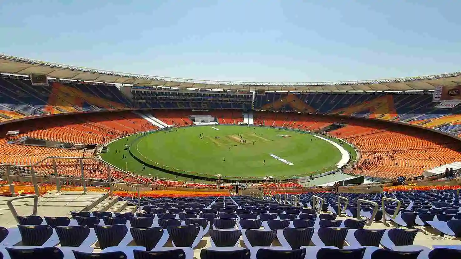 Aerial view of Narendra Modi Stadium in Ahmedabad, India, showing the vast green cricket pitch surrounded by iconic orange and blue seating stands under a clear blue sky — venue for the IND vs NZ T20 World Cup 2026 Final