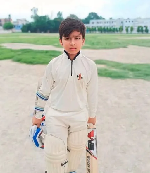 Young Vaibhav Sooryavanshi stands in white cricket kit holding a bat and gloves on a local ground.