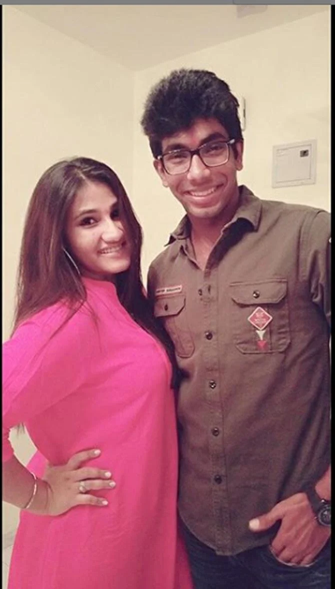 Young Indian cricketer wearing glasses and a brown shirt posing indoors with a female family member dressed in pink.