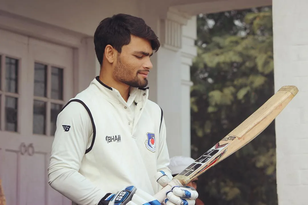 Rinku Singh wearing a white domestic cricket uniform while examining his bat before a match.