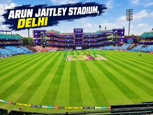 Wide-angle view of Arun Jaitley Stadium in Delhi, showing the green cricket outfield, central pitch, colorful spectator stands and floodlight towers under a clear blue sky.