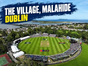 Aerial view of The Village cricket ground in Malahide, Dublin, surrounded by greenery and coastline, with spectators in the stands and the text “The Village, Malahide Dublin.”
