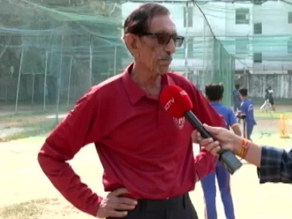 Senior cricket coach in a red shirt speaking during an interview beside outdoor practice nets and young players.