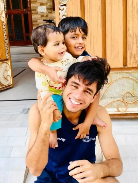 Indian cricketer smiles outdoors while posing closely with two young children in a relaxed off-field photograph.