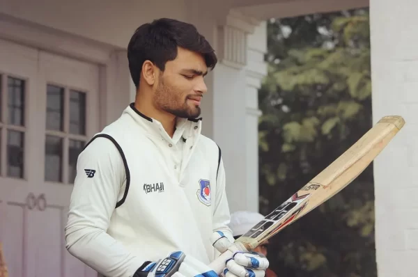 Rinku Singh wearing a white domestic cricket uniform while examining his bat before a match.