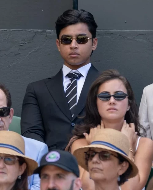Young man in a black suit, striped tie and sunglasses seated among spectators at a cricket venue, used in coverage of Vaibhav Suryavanshi’s rising profile and lifestyle.